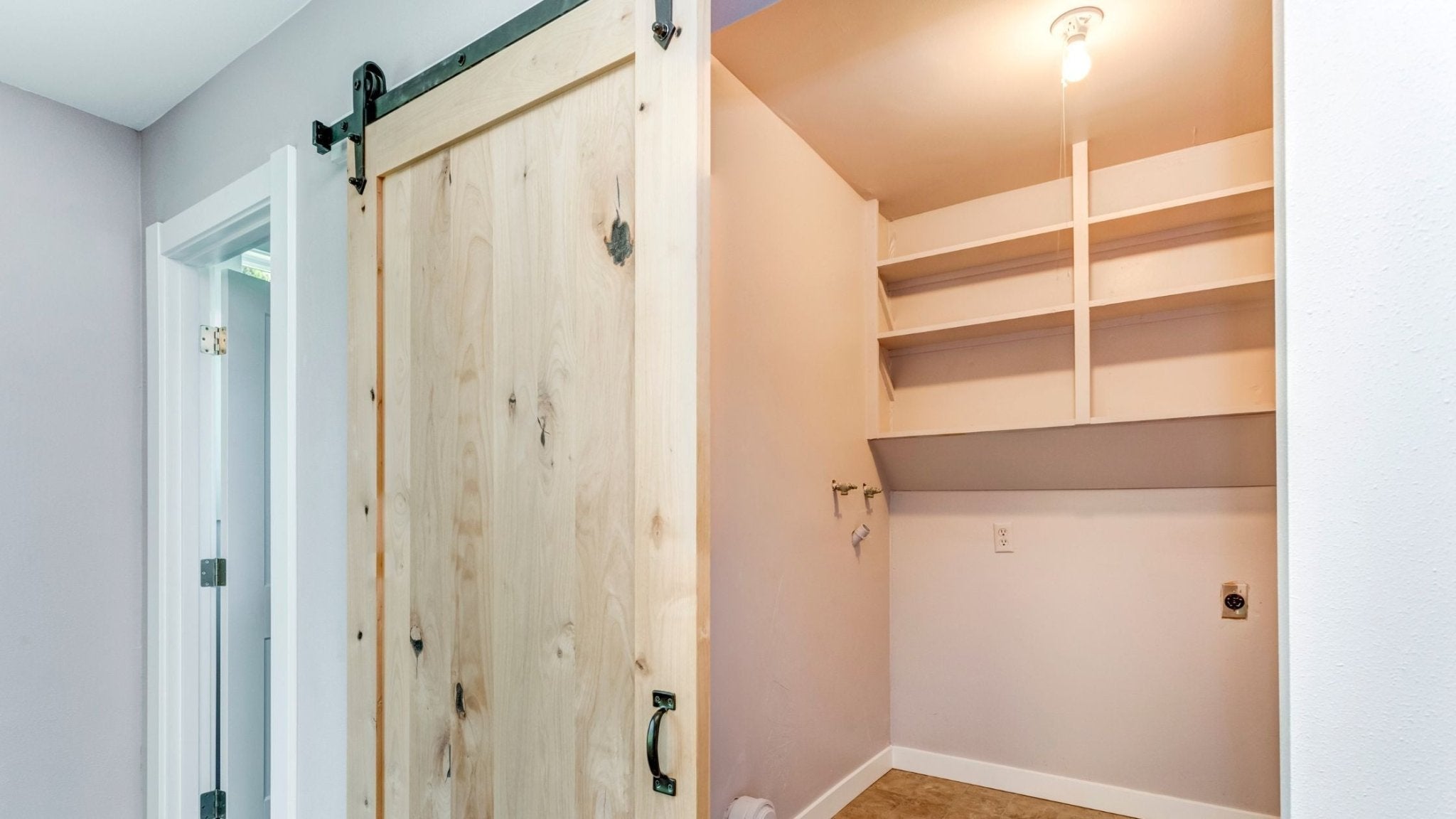 A light wood barn door with visible knots and a dark pull handle, hung on black sliding hardware, partially covers a small closet or utility space with bare shelving and light pink walls.