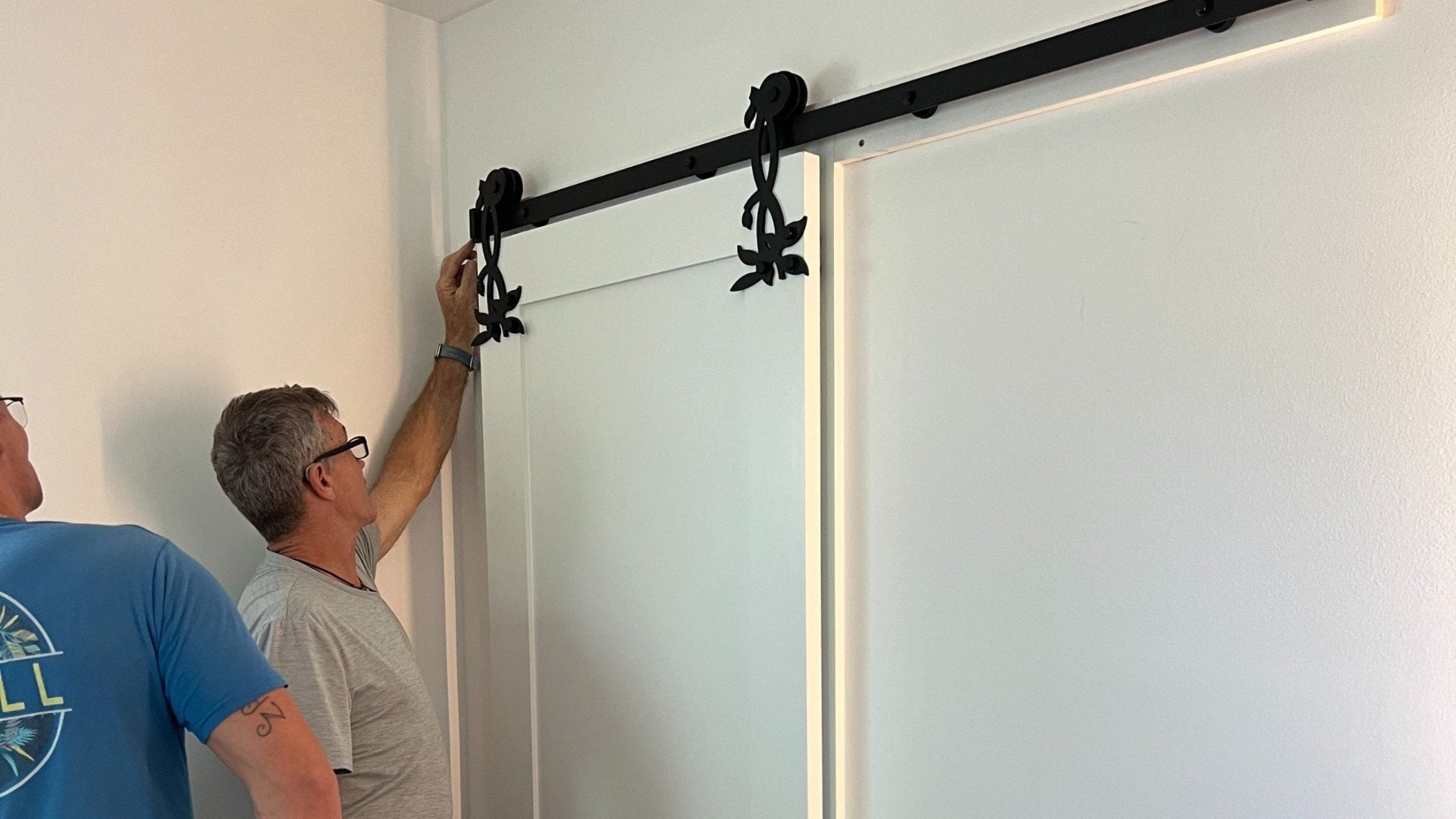 Two men installing a black sliding door track system on a white wall.