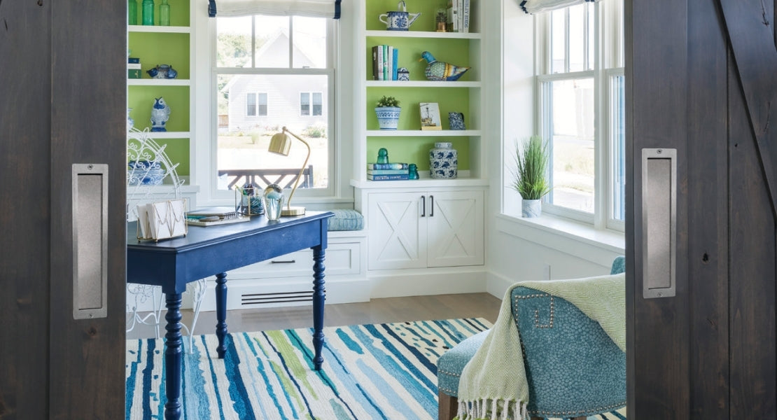 A close-up of a modern home office with two wood barn doors showcasing brushed steel flush pulls. The doors are partially open, revealing a work space with a blue desk, green accent walls, and a colorful striped rug.