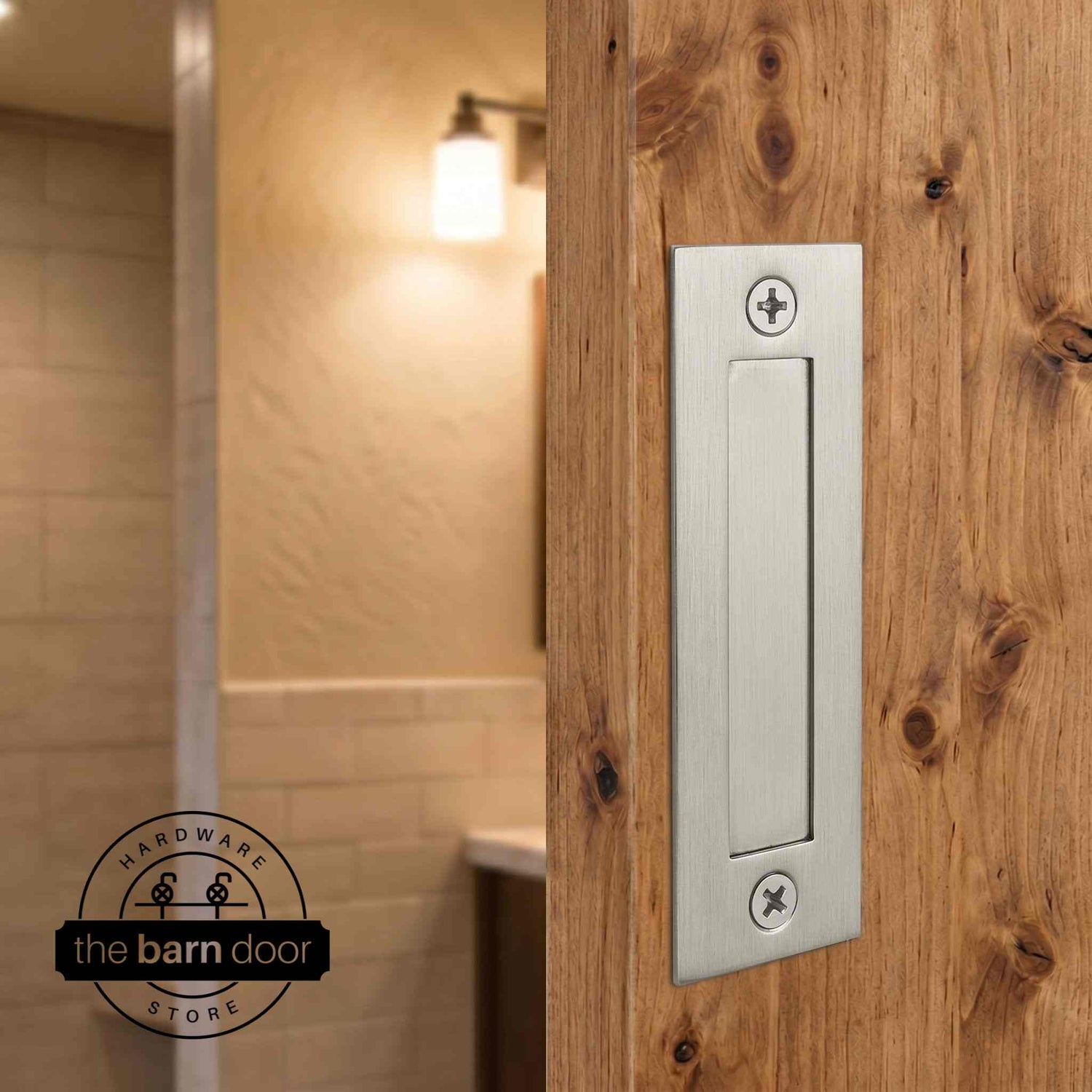 Brushed nickel flush barn door handle mounted on a knotty alder wood door with a warm bathroom interior in the background. By The Barn Door Hardware Store.