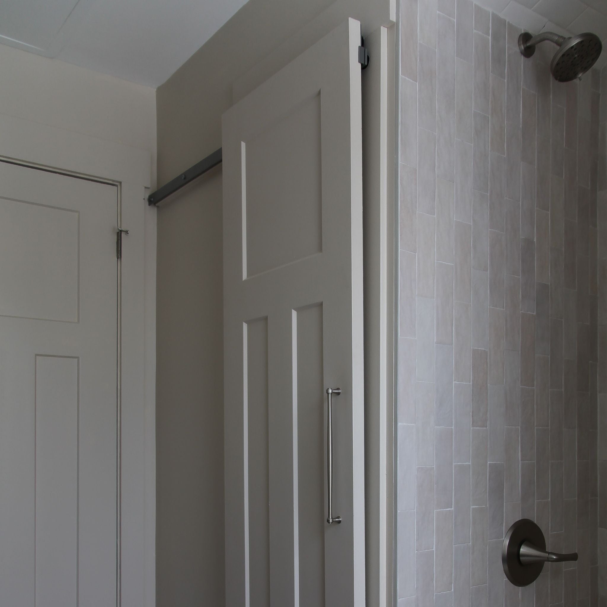 White interior barn door with a vertical pull handle, mounted on a hidden roller track. The door is shown next to a standard swinging closet door on the left and a light grey tiled shower wall with a shower handle on the right.