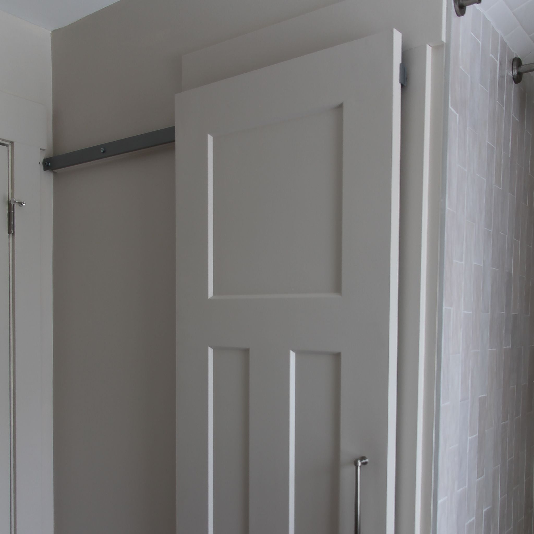 Close-up of the upper half of a light grey-painted interior barn door with four recessed panels, mounted on a hidden roller hardware track. The door slides to the right, partially revealing a light grey wall and an adjacent white standard door frame on the left.