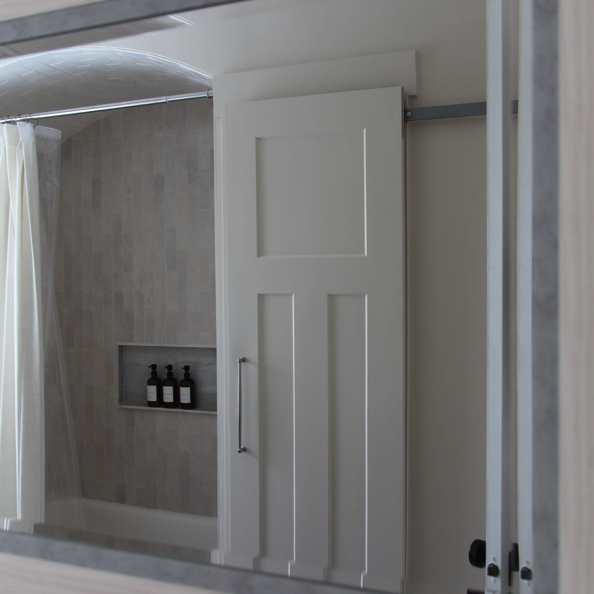 A white paneled interior barn door on a hidden roller track, viewed through a bathroom mirror. The door is positioned in front of a shower/tub area with light grey vertical subway tile, a recessed niche with bottles, and a shower curtain.