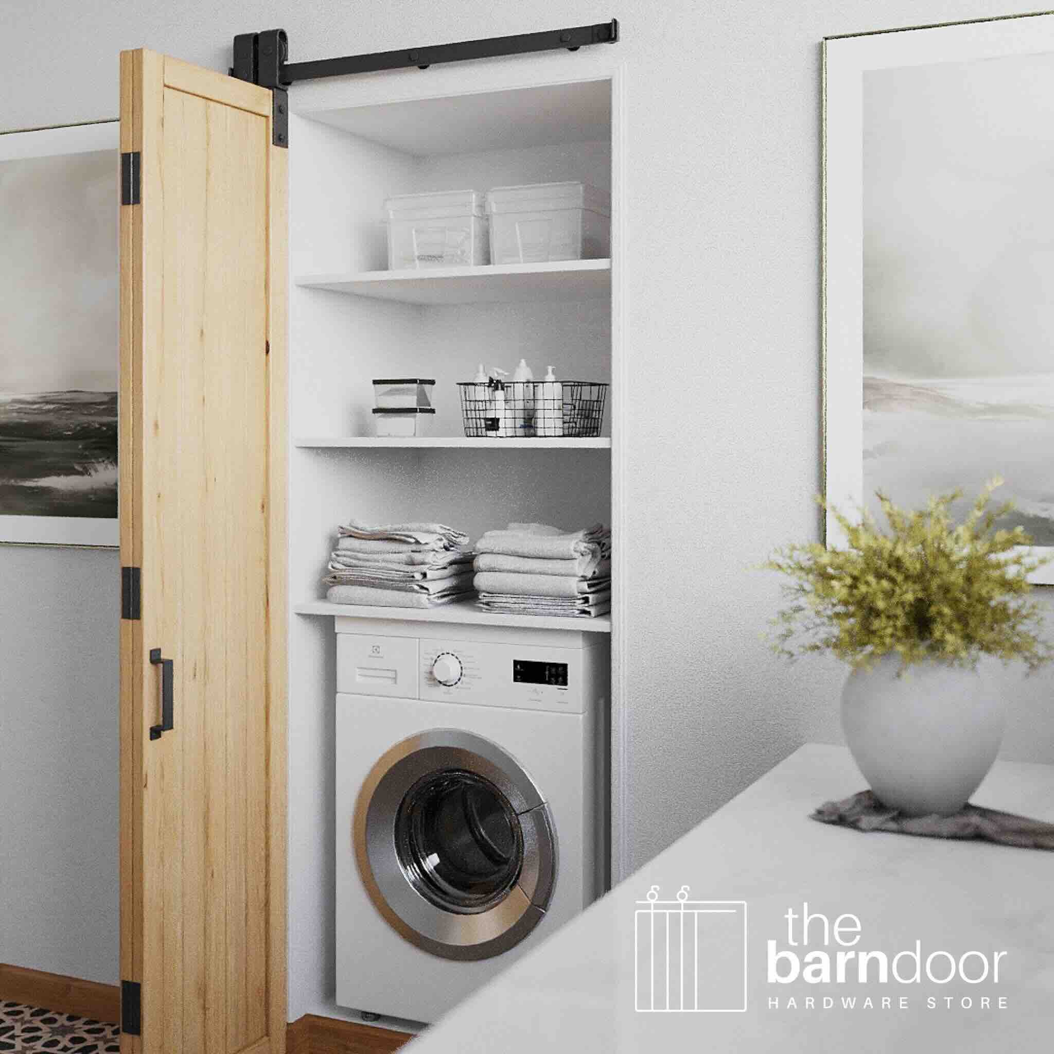 Wooden bifold barn door opening to reveal a laundry room with a washing machine, shelves, and decor.