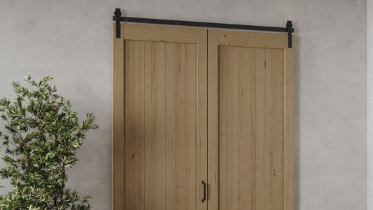  Close-up of two light natural wood bifold barn door panels with vertical plank construction, installed on a black top-mount sliding hardware track against a gray wall. 