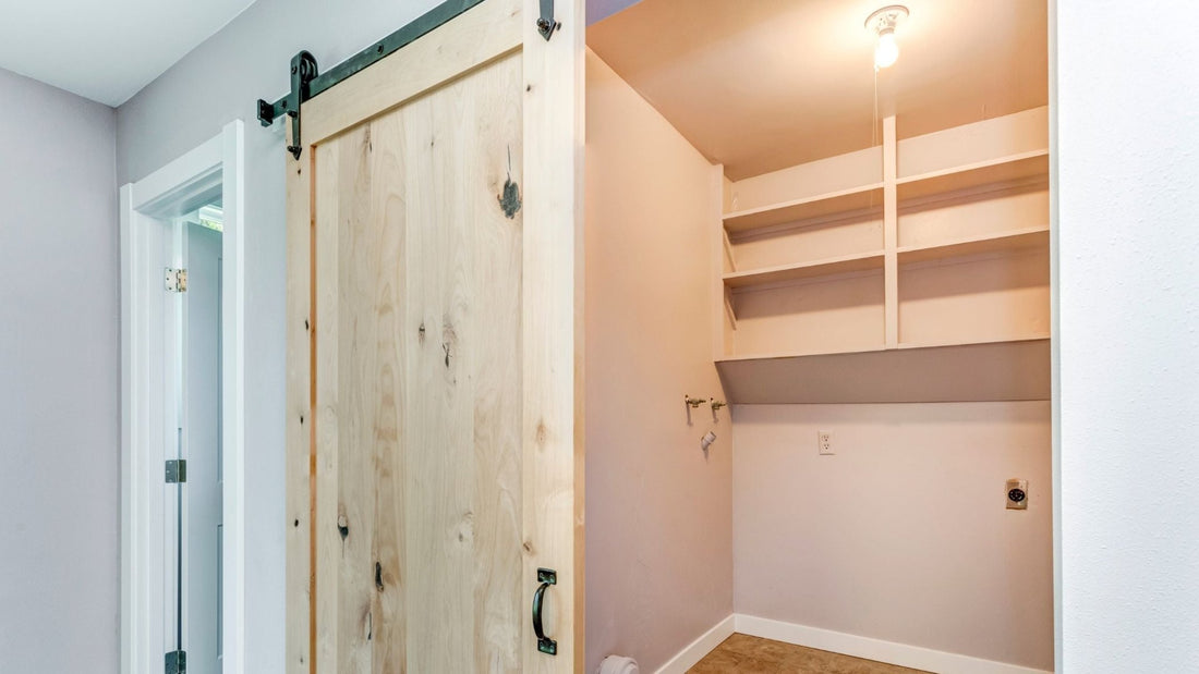 A light wood barn door with visible knots and a dark pull handle, hung on black sliding hardware, partially covers a small closet or utility space with bare shelving and light pink walls.