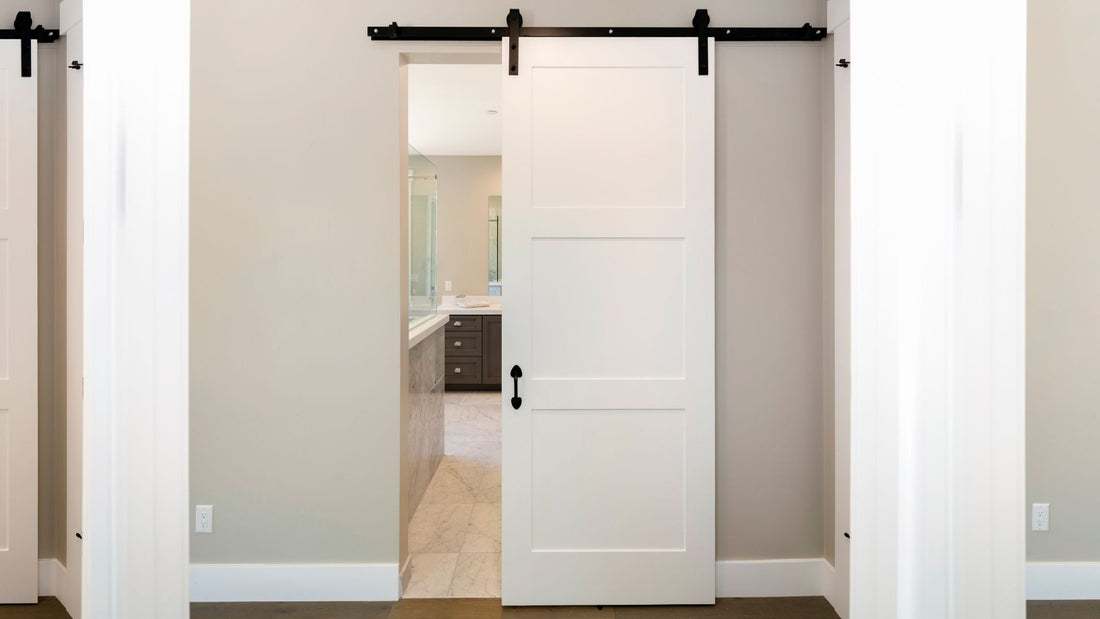A white three-panel sliding barn door, slightly ajar, installed with black sliding hardware, revealing a modern bathroom with marble floors and dark wood vanity inside.