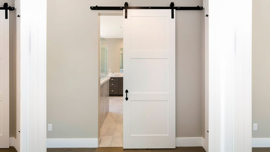 A white three-panel sliding barn door, slightly ajar, installed with black sliding hardware, revealing a modern bathroom with marble floors and dark wood vanity inside.