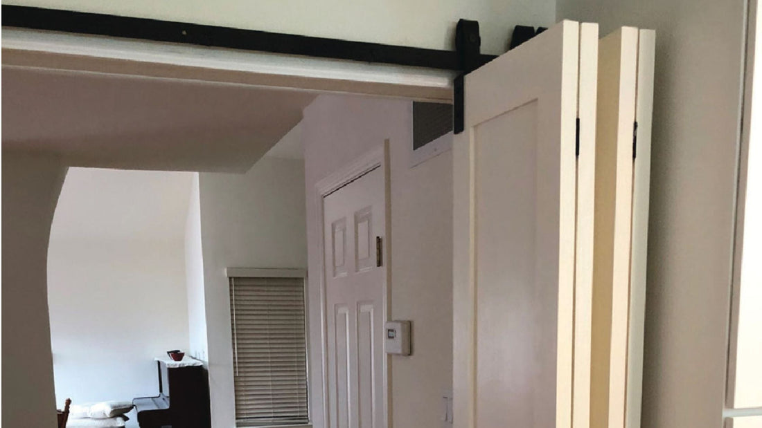 Close-up of white bifold barn door panels stacked open, installed with black sliding hardware over an interior doorway, revealing a room with a window and a white standard door inside.