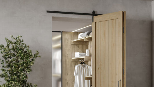 A modern light wood barn door with black bifold sliding hardware partially open, revealing a well-lit, organized walk-in closet with wood shelves and clothing inside.