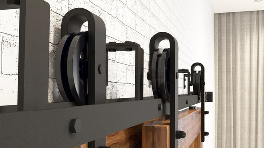 Close-up of matte black steel bypass barn door hardware, featuring large dual roller hangers and a rail mounted on a white painted brick wall. The wooden door surface is partially visible beneath the rail.