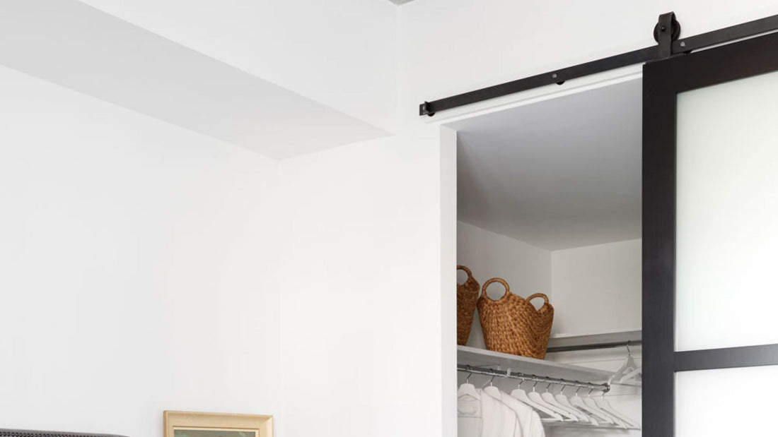 Modern closet with a black-framed barn door and opaque white panel. Black sliding hardware is mounted above the opening. Inside, two woven baskets rest on a shelf above white hangers.
