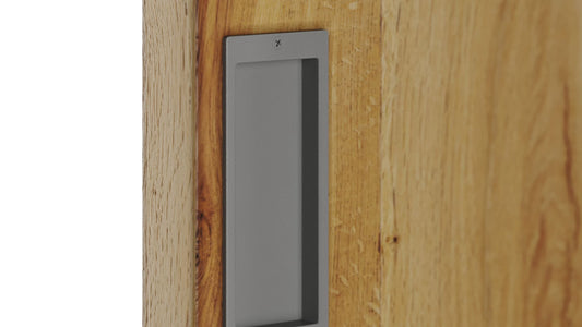Close-up of a rectangular, matte gray flush pull handle recessed into a natural, light oak wood barn door.