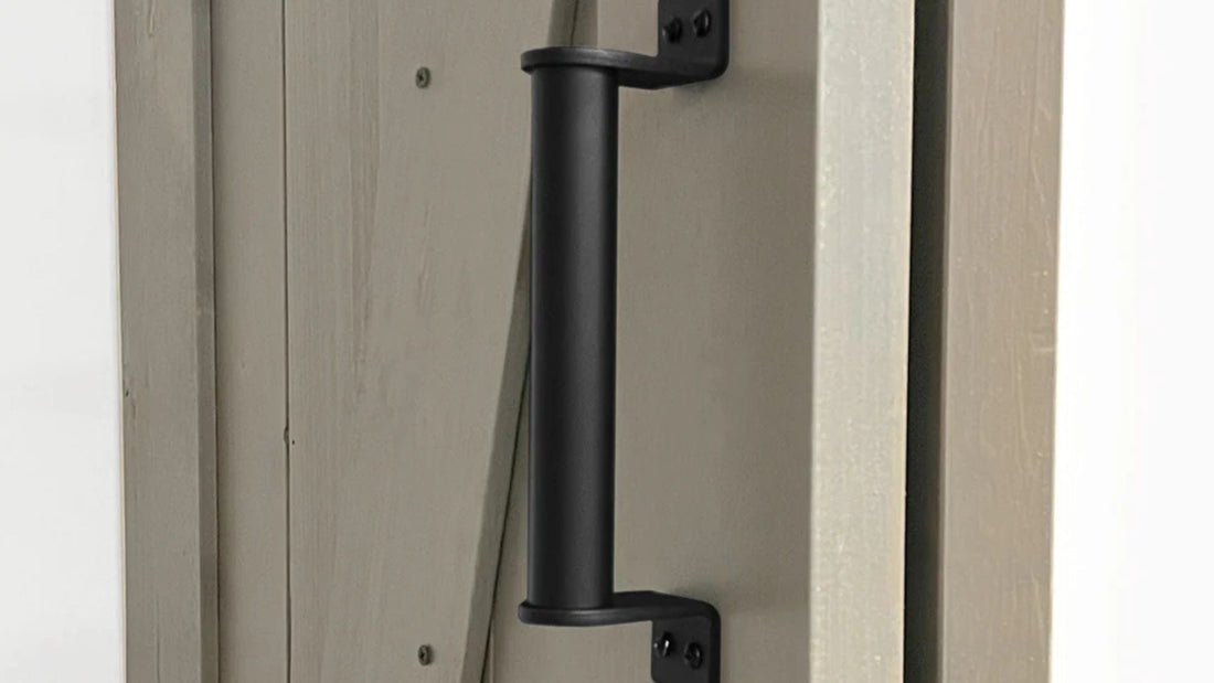 Close-up of a sturdy, cylindrical matte black barn door pull handle mounted with angled brackets on a door panel painted a light grayish-taupe color.