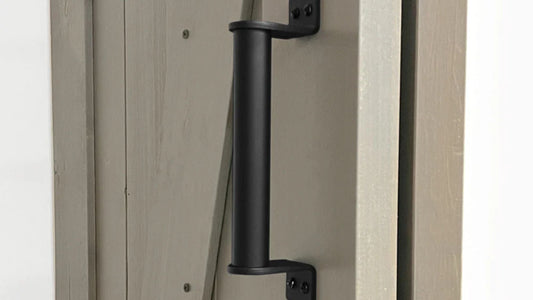 Close-up of a sturdy, cylindrical matte black barn door pull handle mounted with angled brackets on a door panel painted a light grayish-taupe color.