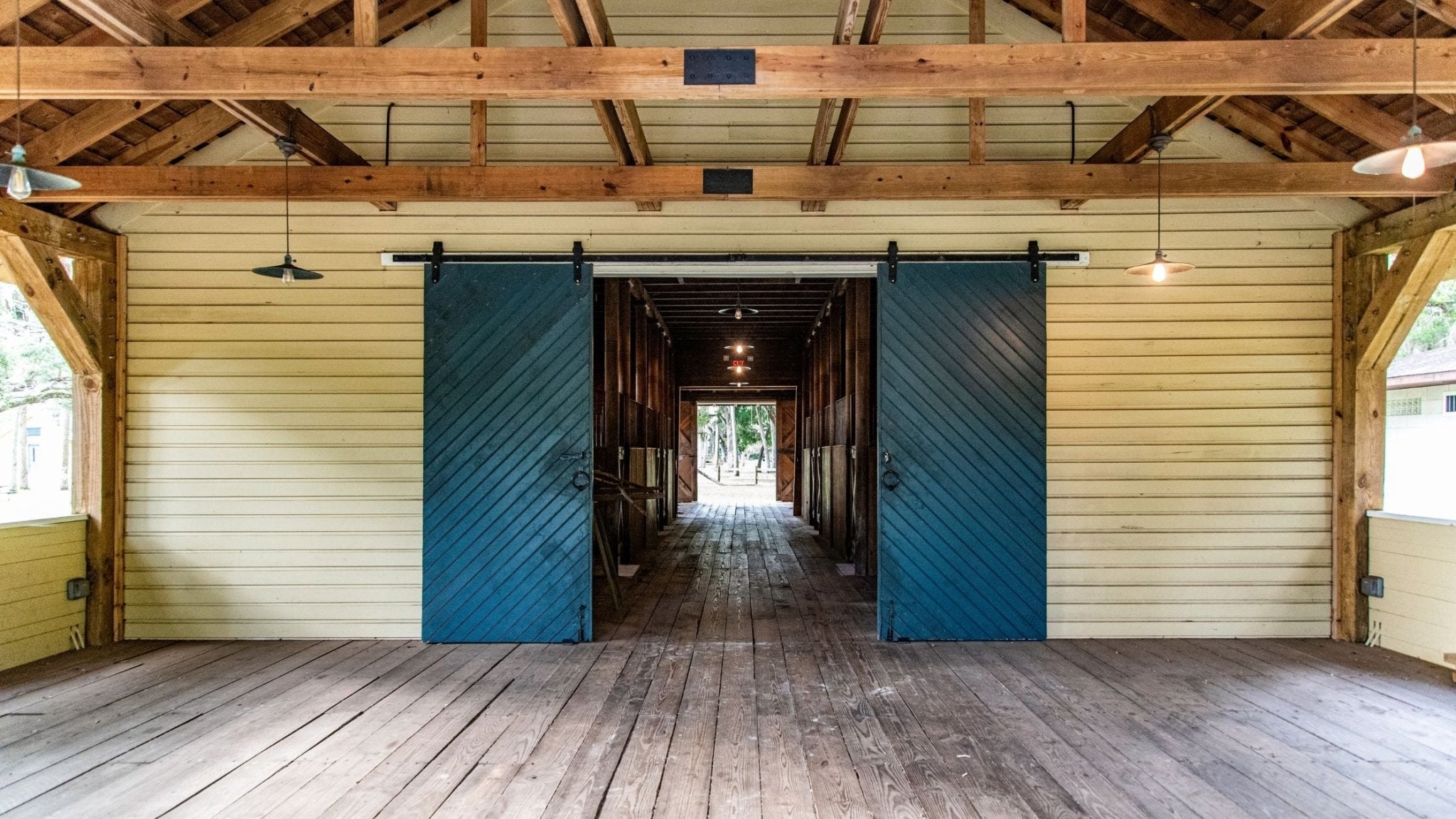 Two large teal barn doors with a diagonal plank design, installed on a black top-mount sliding track, slightly open to reveal a hallway inside a rustic barn.