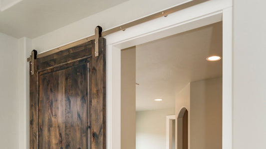 Dark stained wood sliding barn door with knotty texture, installed with bright brushed metal J-strap hardware and track above a white-framed doorway.