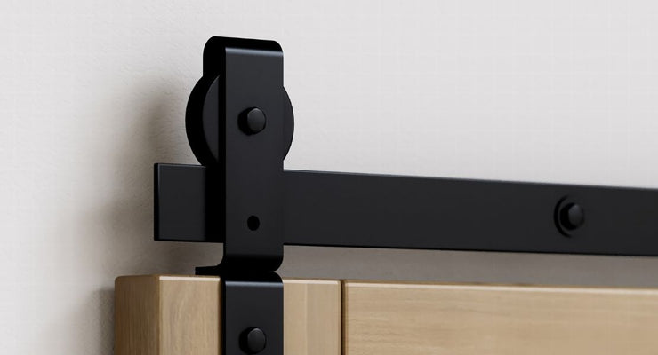 A close-up detail shot of black industrial-style J-strap roller hardware mounted on the top edge of a light wood bifold door.