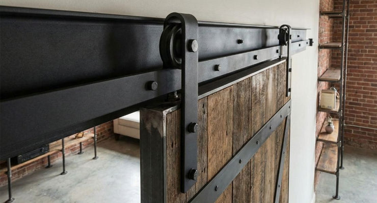 A close-up view of heavy-duty industrial barn door hardware featuring large black steel rollers and exposed bolts. The hardware holds a rugged, reclaimed wood door reinforced with metal strapping.