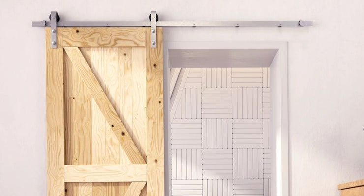 A light unfinished wood barn door with a K-brace design mounted on a sleek silver stainless steel track system in a modern white room.