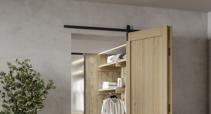 A modern, light-wood bifold barn door mounted on a black track, partially open to reveal a well-lit closet with wooden shelving.
