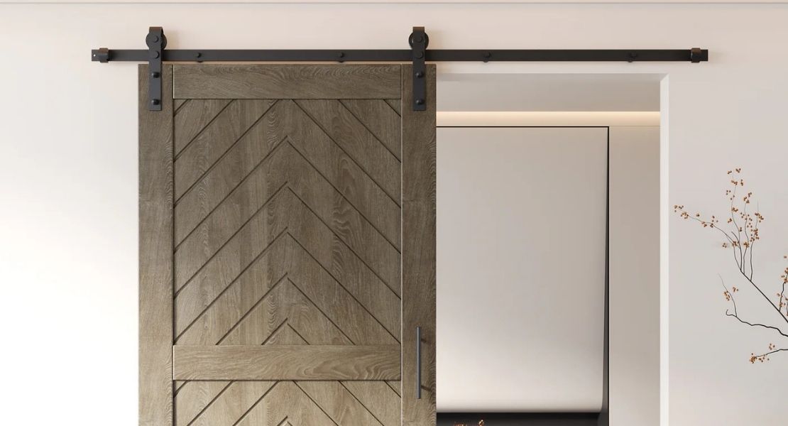 A chic modern barn door featuring a chevron wood grain pattern in a grey-brown finish, hanging on a black track in a minimalist white room.