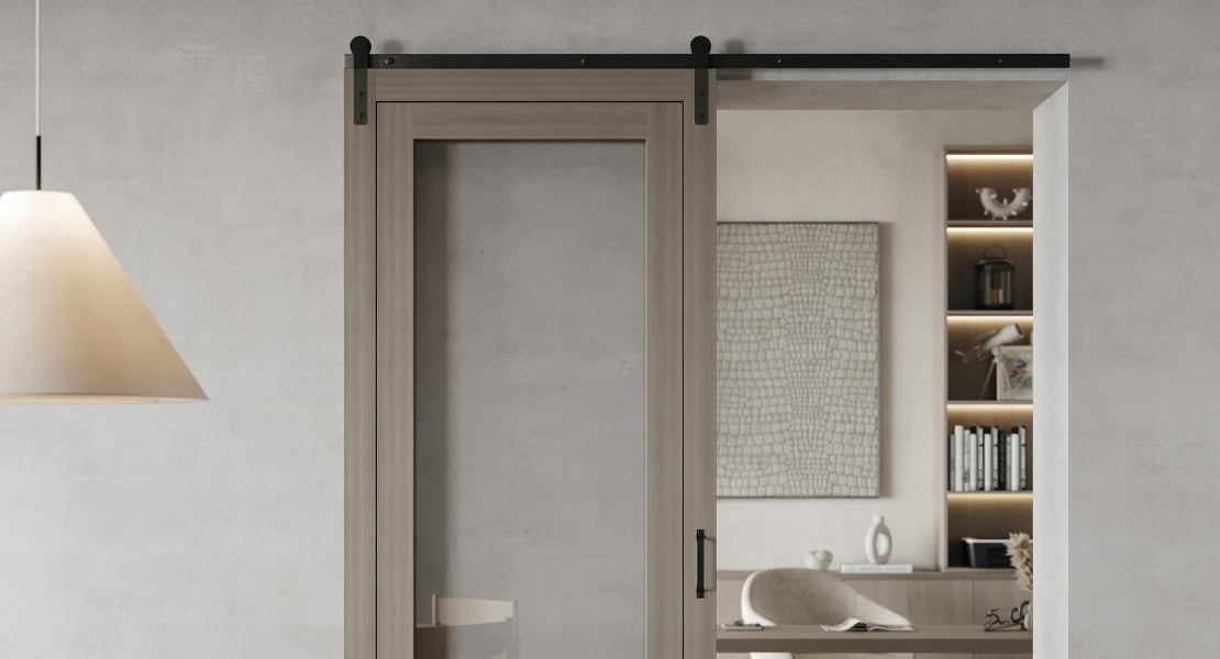 A classic J-strap style single track sliding barn door hardware kit in a matte black finish, installed on a light-colored wood door with a single glass panel.