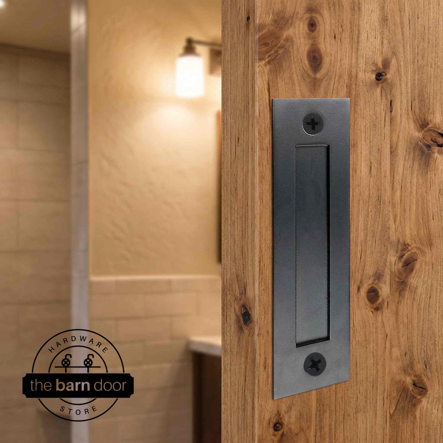 Matte black flush barn door handle mounted on a knotty alder wood door with a warm bathroom interior in the background. By The Barn Door Hardware Store.