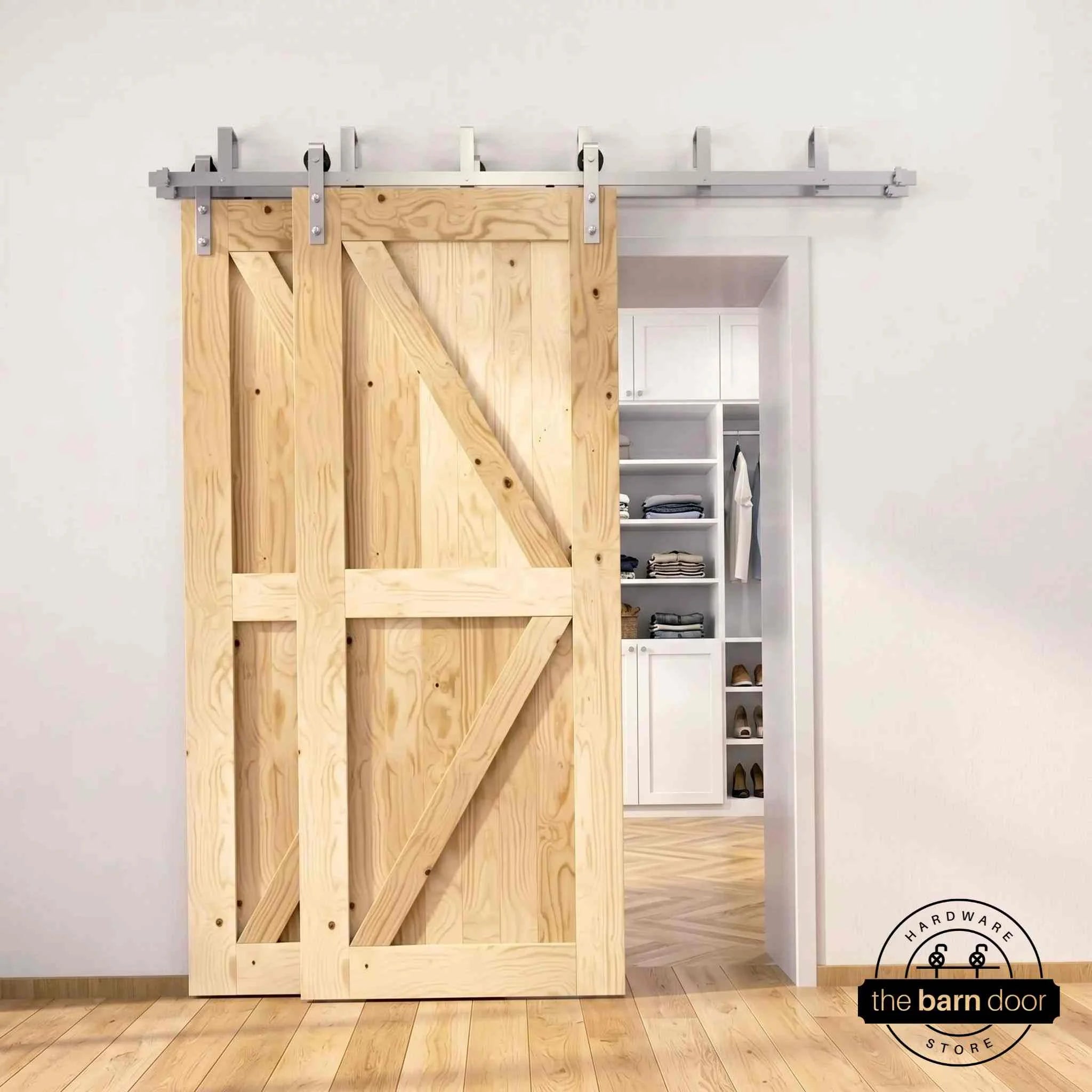 Brushed nickel double bypass barn door kit with two unfinished K-brace doors mounted on a dual-rail track, partially open to reveal a white built-in closet with folded clothing, hanging garments, and shoe shelving, set against a white wall with light herringbone and oak plank flooring.