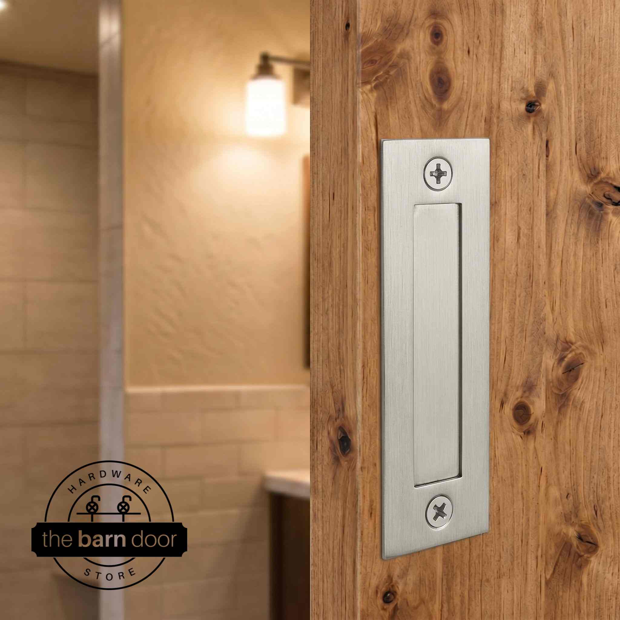 Brushed nickel flush barn door handle mounted on a knotty alder wood door with a warm bathroom interior in the background. By The Barn Door Hardware Store.