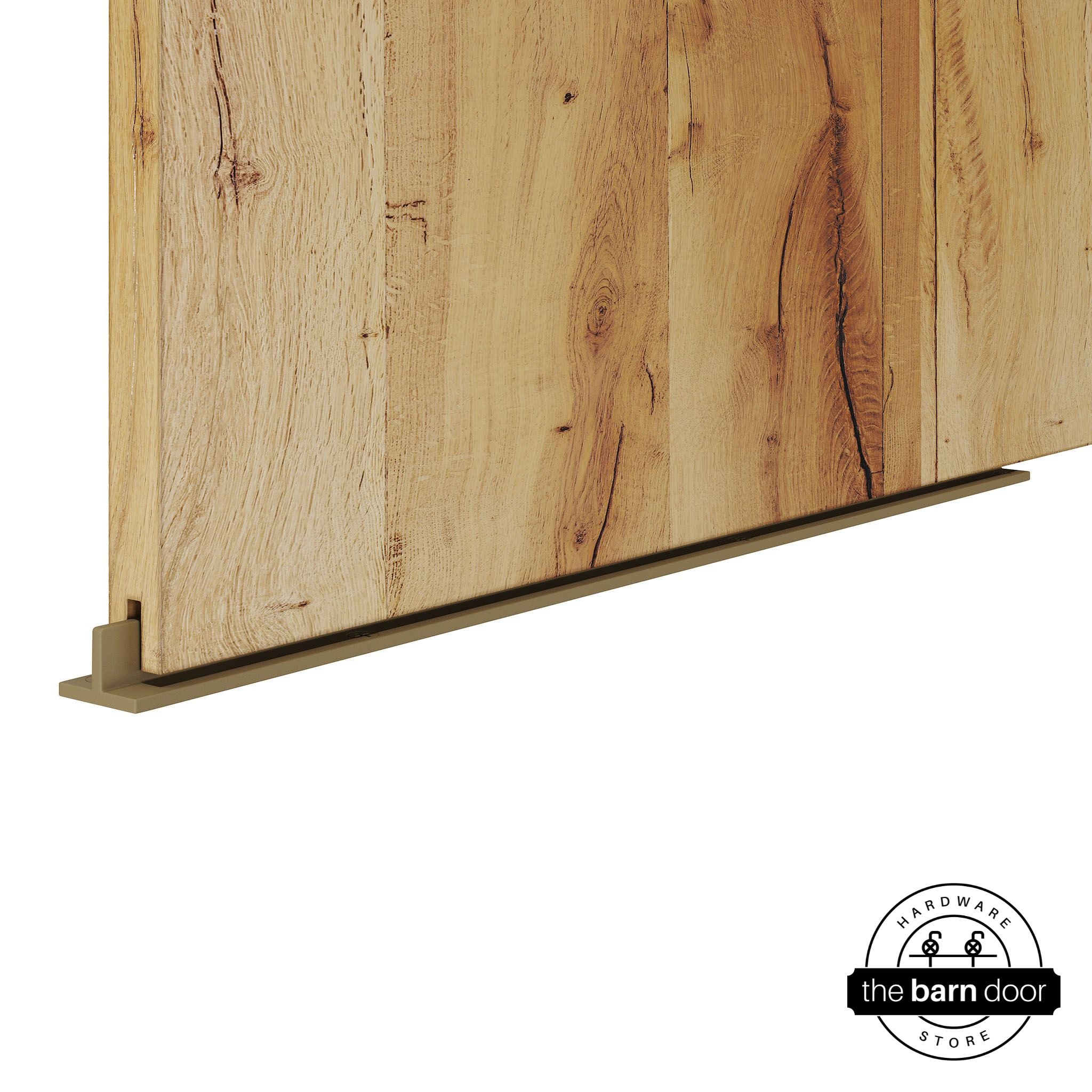 Gold continuous T-guide floor track for barn doors by The Barn Door Hardware Store. Close-up of a heavy-duty bottom floor guide installed on a rustic wood door for stabilization and smooth sliding operation.