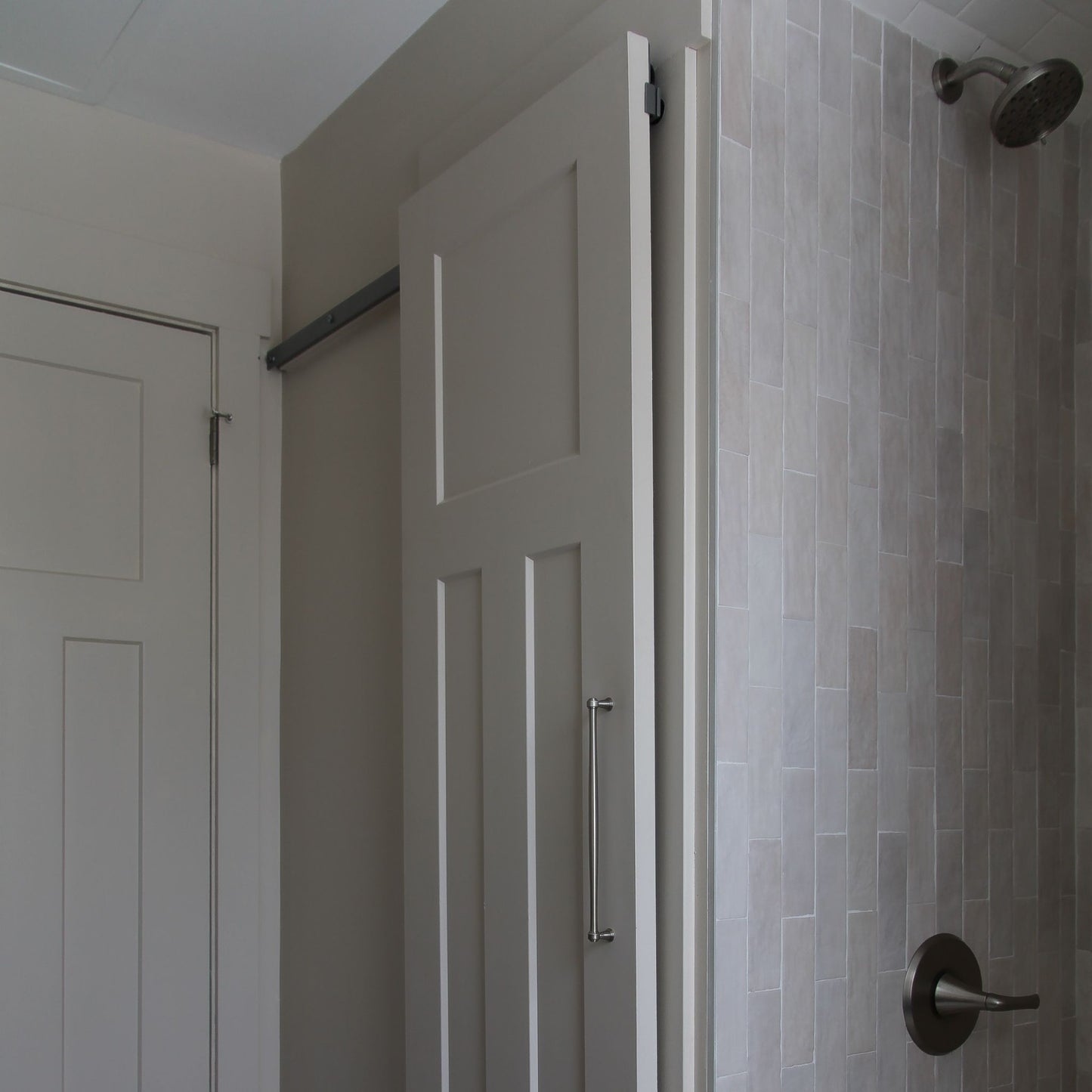 White interior barn door with a vertical pull handle, mounted on a hidden roller track. The door is shown next to a standard swinging closet door on the left and a light grey tiled shower wall with a shower handle on the right.