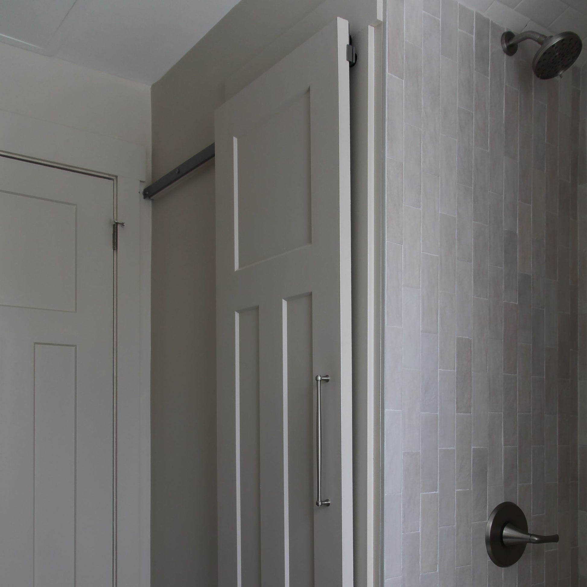 White interior barn door with a vertical pull handle, mounted on a hidden roller track. The door is shown next to a standard swinging closet door on the left and a light grey tiled shower wall with a shower handle on the right.