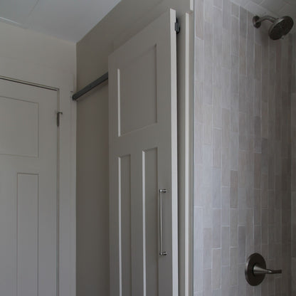 White interior barn door with a vertical pull handle, mounted on a hidden roller track. The door is shown next to a standard swinging closet door on the left and a light grey tiled shower wall with a shower handle on the right.