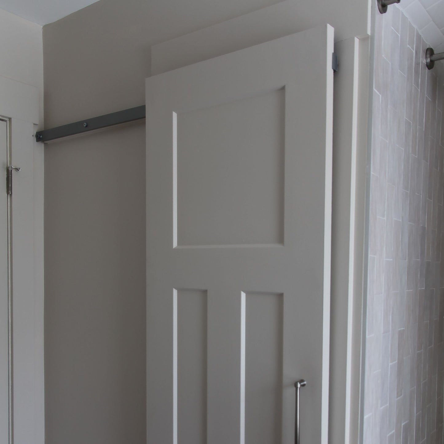 Close-up of the upper half of a light grey-painted interior barn door with four recessed panels, mounted on a hidden roller hardware track. The door slides to the right, partially revealing a light grey wall and an adjacent white standard door frame on the left.