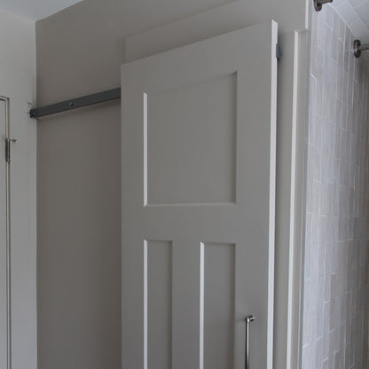 Close-up of the upper half of a light grey-painted interior barn door with four recessed panels, mounted on a hidden roller hardware track. The door slides to the right, partially revealing a light grey wall and an adjacent white standard door frame on the left.