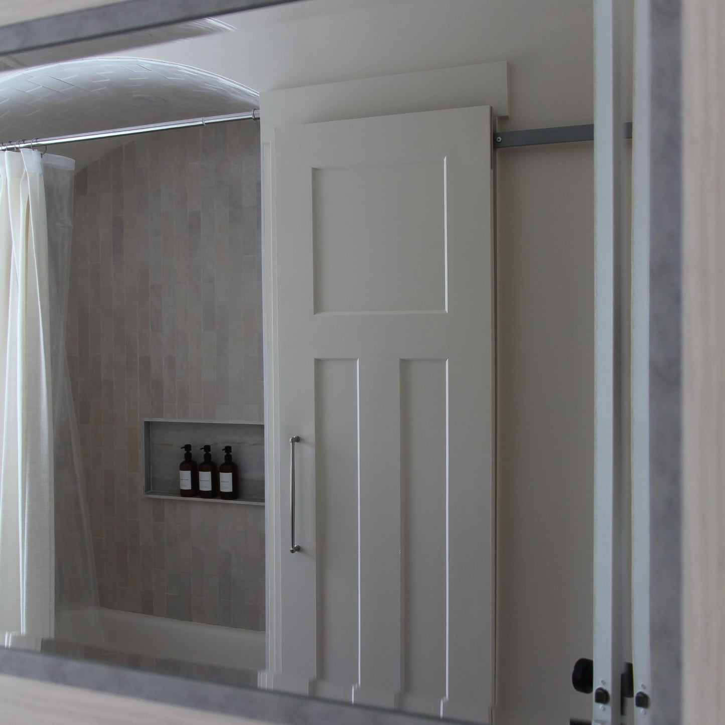 A white paneled interior barn door on a hidden roller track, viewed through a bathroom mirror. The door is positioned in front of a shower/tub area with light grey vertical subway tile, a recessed niche with bottles, and a shower curtain.