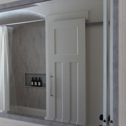 A white paneled interior barn door on a hidden roller track, viewed through a bathroom mirror. The door is positioned in front of a shower/tub area with light grey vertical subway tile, a recessed niche with bottles, and a shower curtain.