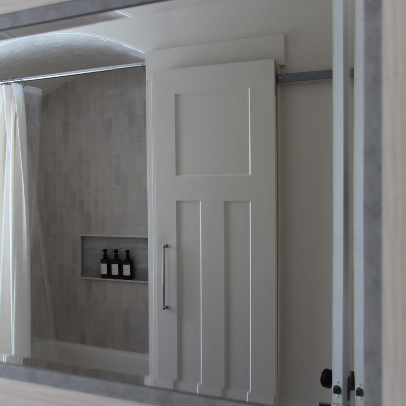 A white paneled interior barn door on a hidden roller track, viewed through a bathroom mirror. The door is positioned in front of a shower/tub area with light grey vertical subway tile, a recessed niche with bottles, and a shower curtain.