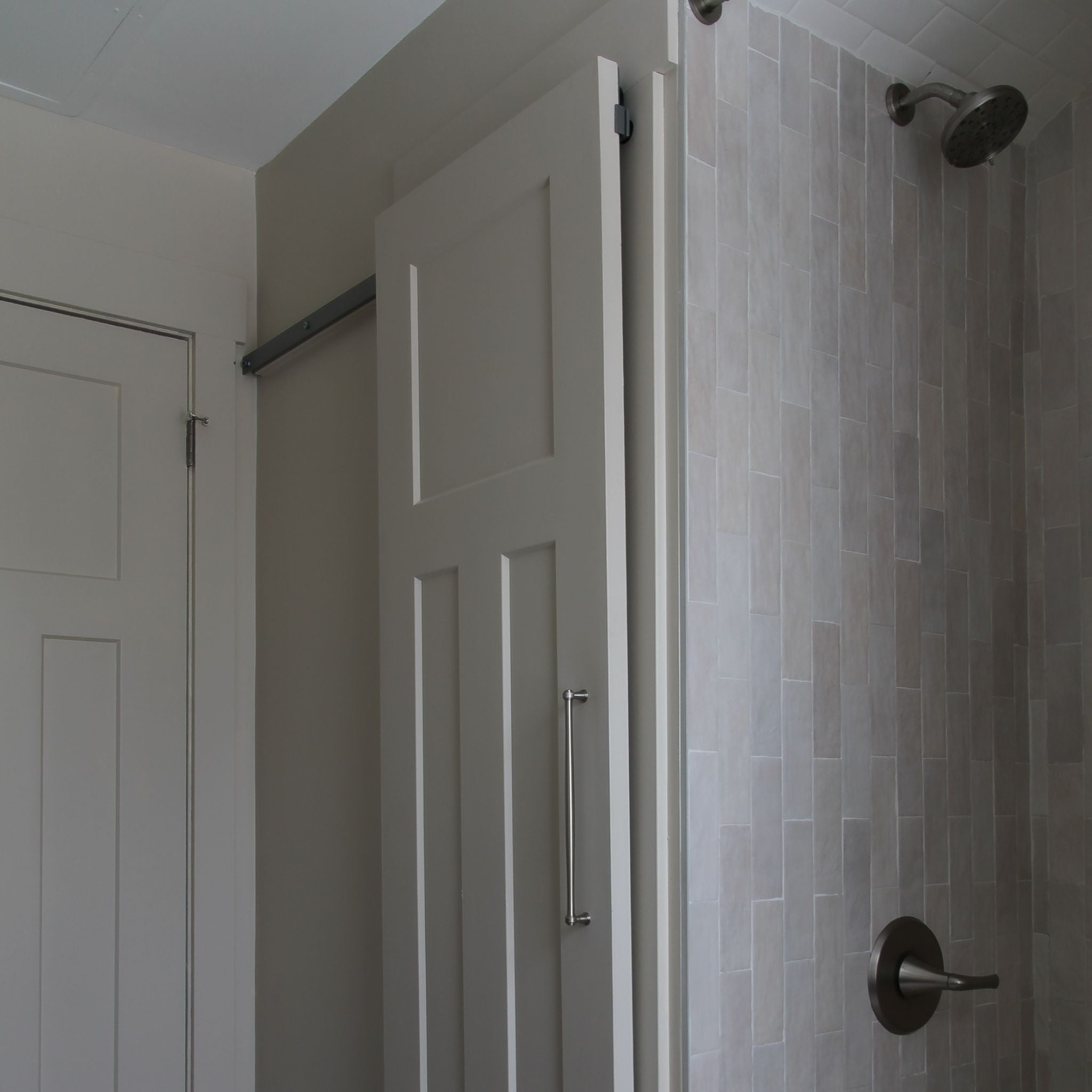 Close-up of a white interior barn door with a vertical pull handle, partially covering a tiled shower area with a rain showerhead. The hidden top roller track is visible.