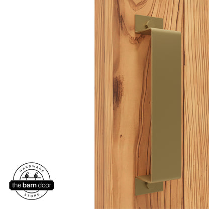 A Jacob's Gold, arch-shaped barn door handle is mounted on a light-colored wooden door with a vertical grain pattern.