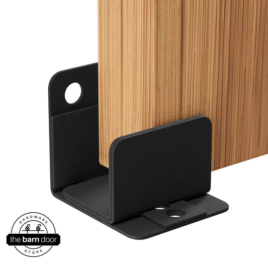 A matte black U-shaped floor guide with a side plate, holding a light wood barn door in place.