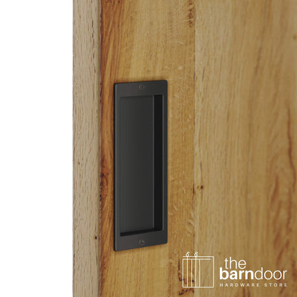 A close-up shot of a recessed rectangular flush pull handle in a matte black finish, installed on a light brown wooden barn door.