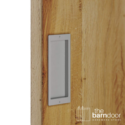 A close-up shot of a recessed rectangular flush pull handle in a PC Chrome finish, installed on a light brown wooden barn door.