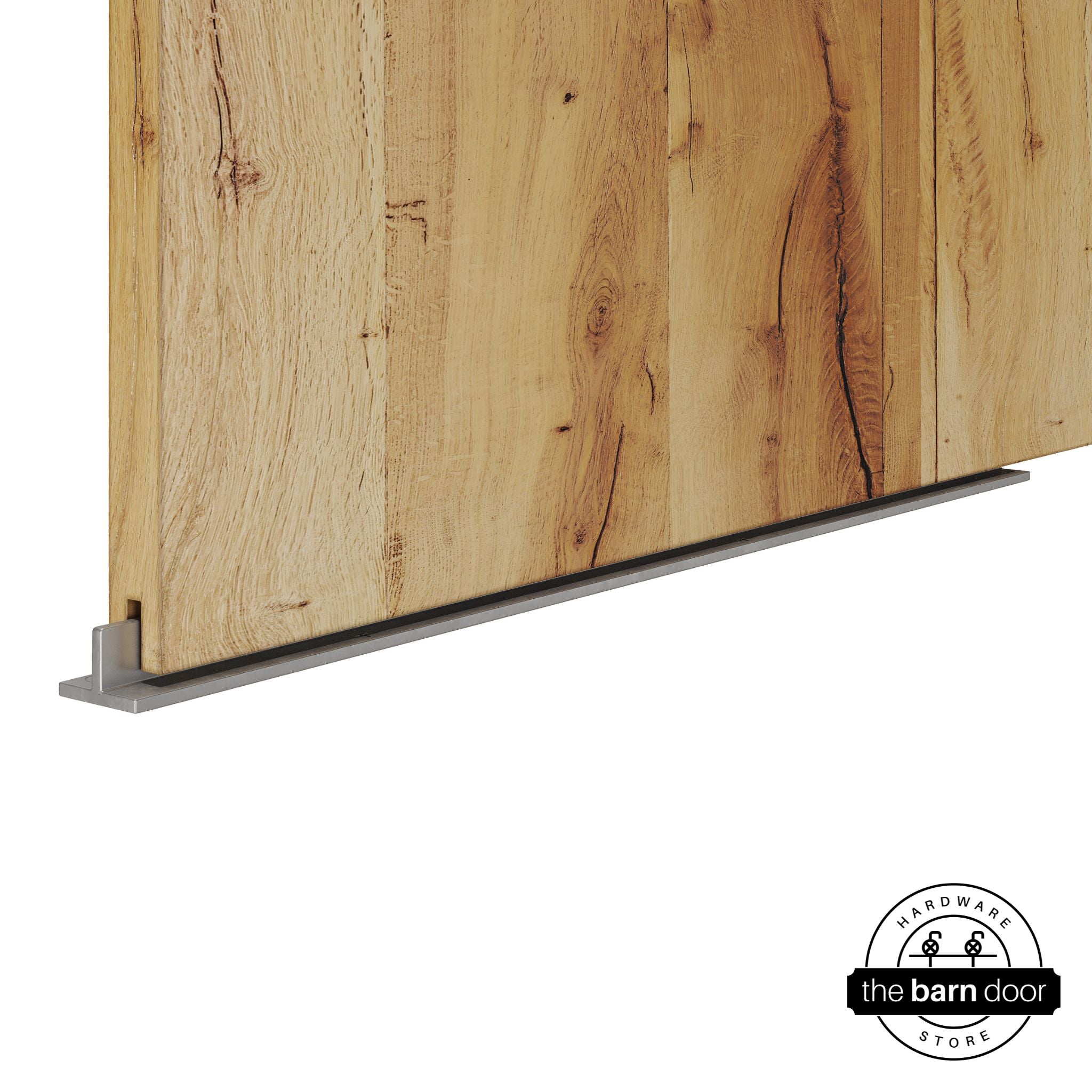 Raw steel continuous T-guide floor track for barn doors by The Barn Door Hardware Store. Close-up of a heavy-duty bottom floor guide installed on a rustic wood door for stabilization and smooth sliding operation.