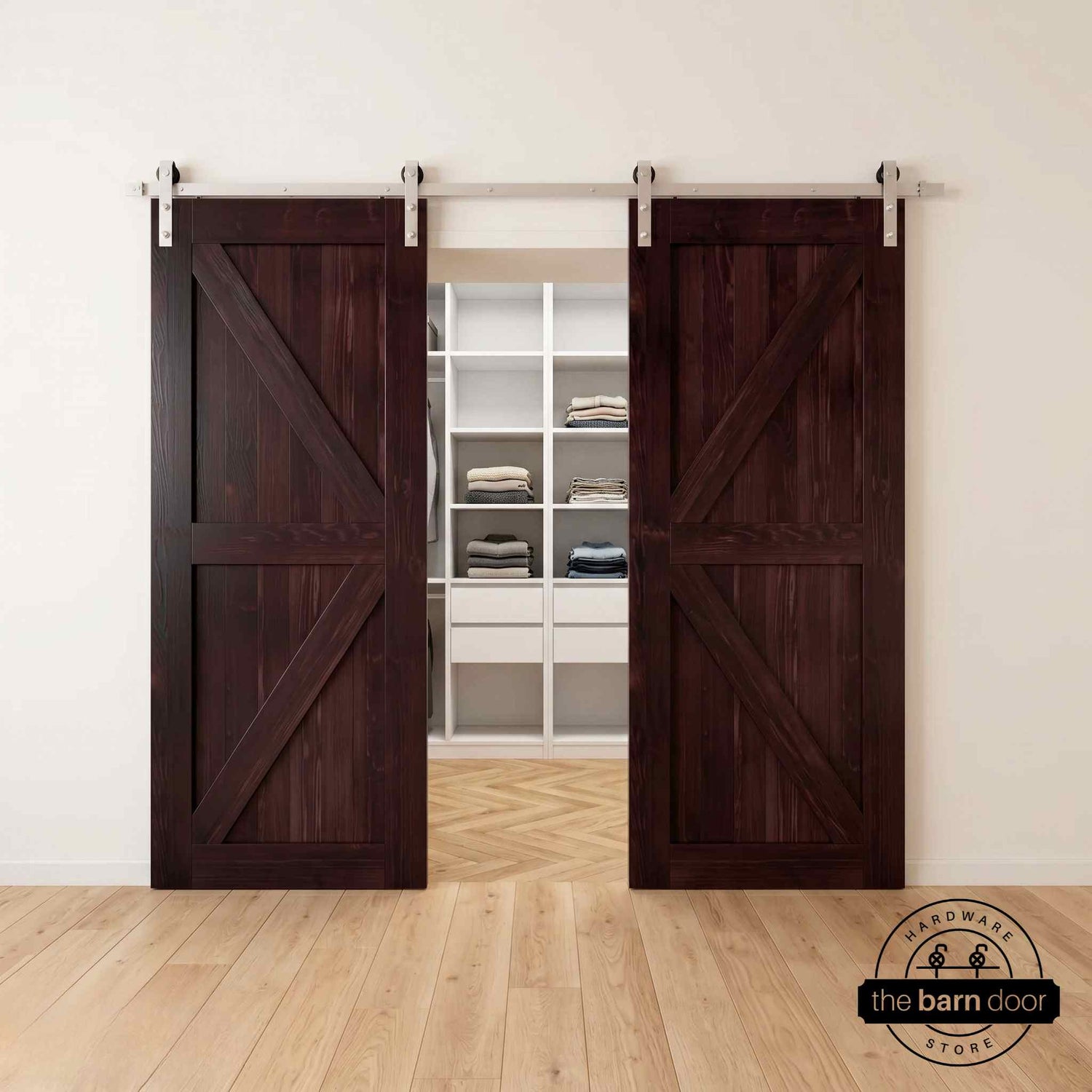 Two wood double barn doors with a two-panel vertical plank design in a rich, dark Red Mahogany stain, installed with brushed nickel sliding hardware, partially revealing a living room area.