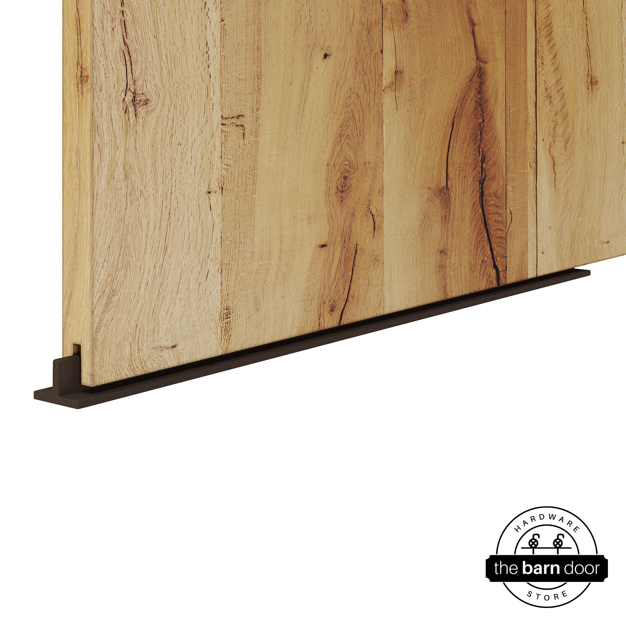 Rustic Brown continuous T-guide floor track for barn doors by The Barn Door Hardware Store. Close-up of a heavy-duty bottom floor guide installed on a rustic wood door for stabilization and smooth sliding operation.