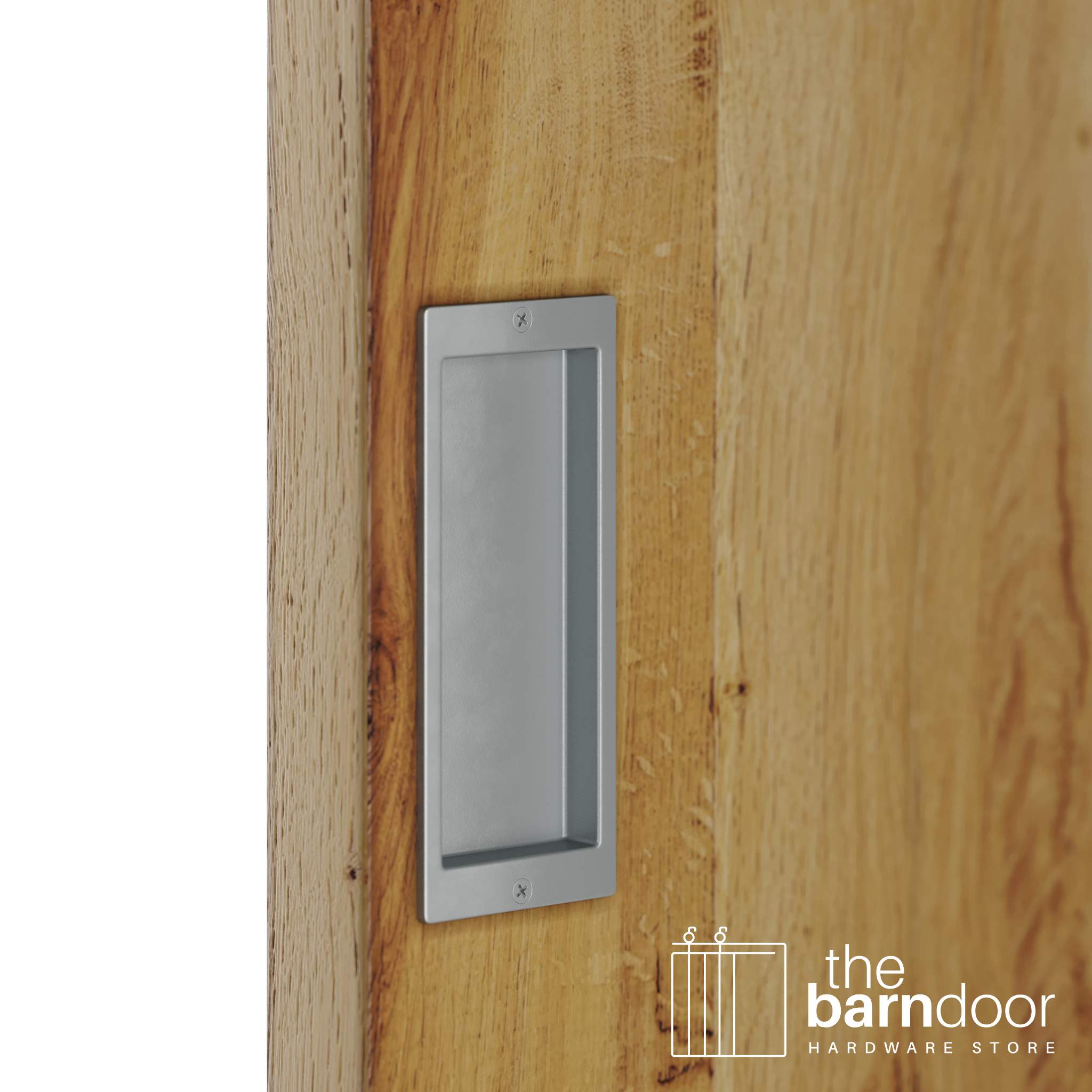 A close-up shot of a recessed rectangular flush pull handle in a silver metallic finish, installed on a light brown wooden barn door.