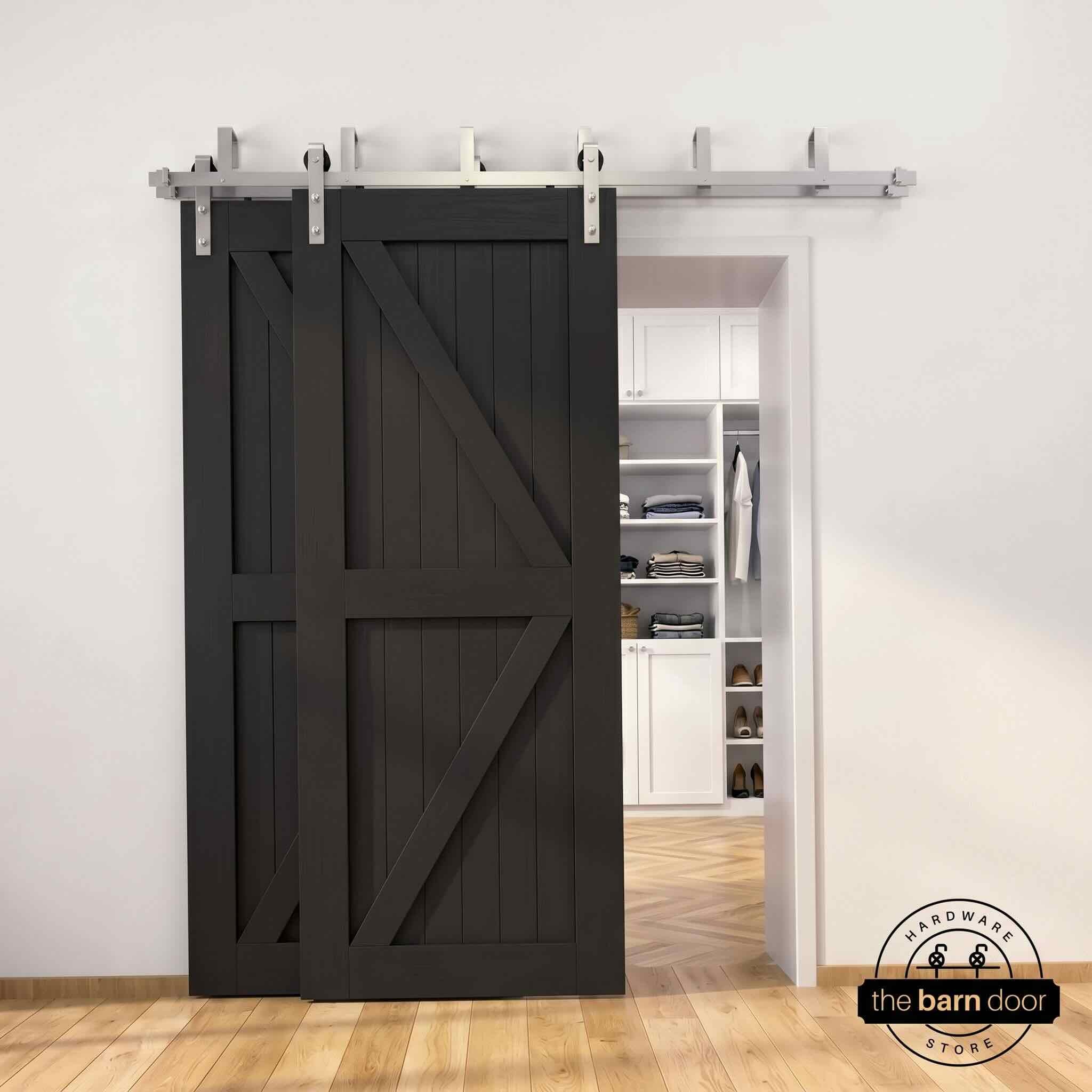 Brushed nickel double bypass barn door kit with two black K-brace doors mounted on a dual-rail track, partially open to reveal a white built-in closet with folded clothing, hanging garments, and shoe shelving, set against a white wall with light herringbone and oak plank flooring.