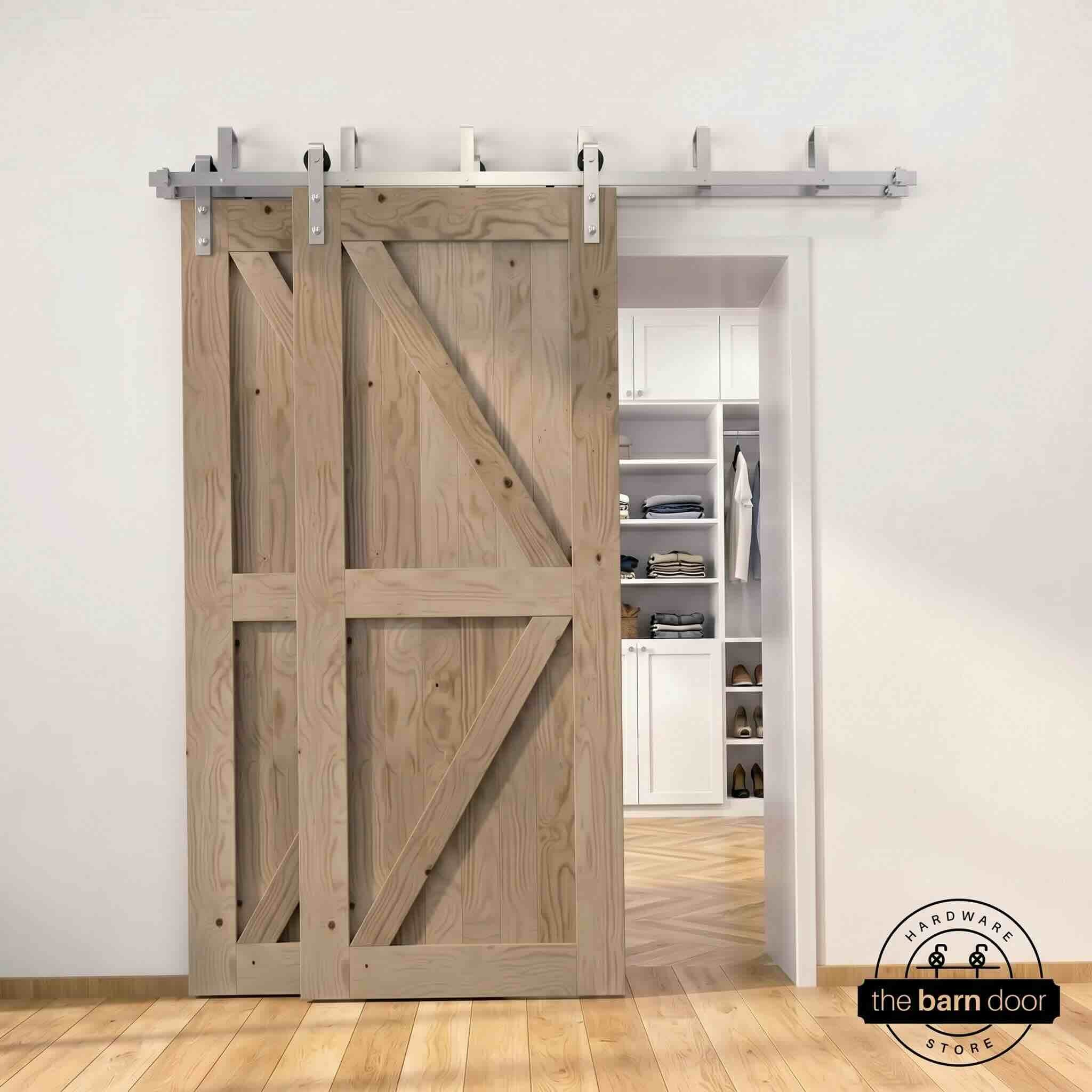 Brushed nickel double bypass barn door kit with two classic gray K-brace doors mounted on a dual-rail track, partially open to reveal a white built-in closet with folded clothing, hanging garments, and shoe shelving, set against a white wall with light herringbone and oak plank flooring.